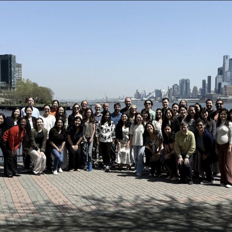 A large, diverse group of people posing for a group photo on a waterfront promenade with a city skyline and river in the background.