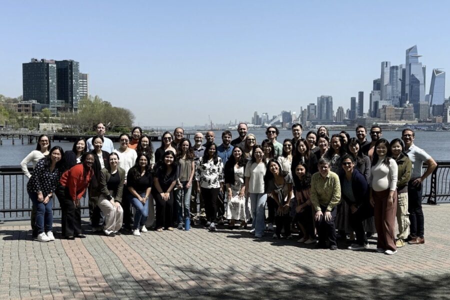 A large, diverse group of people posing for a group photo on a waterfront promenade with a city skyline and river in the background.
