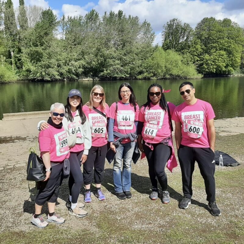 • Six Genesis Research Group team members posing in pink t-shirts by a lake during a breast cancer charity walk.