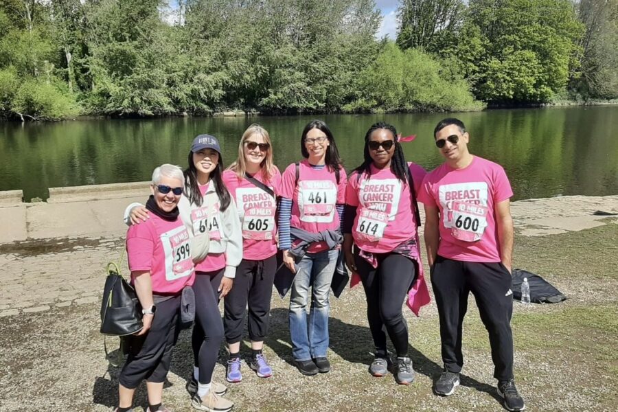 • Six Genesis Research Group team members posing in pink t-shirts by a lake during a breast cancer charity walk.